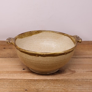 May include: A handcrafted ceramic bowl with a speckled beige interior and a dark brown rim. The bowl has two decorative handles and is placed on a wooden surface. The bowl is a neutral colour and suitable for serving food.