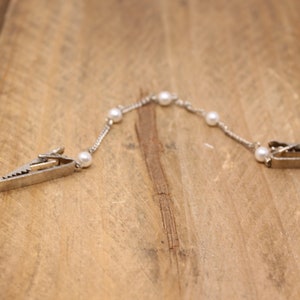May include: A silver chain with white pearl accents and two silver clips on each end. The chain is lying on a wooden surface.