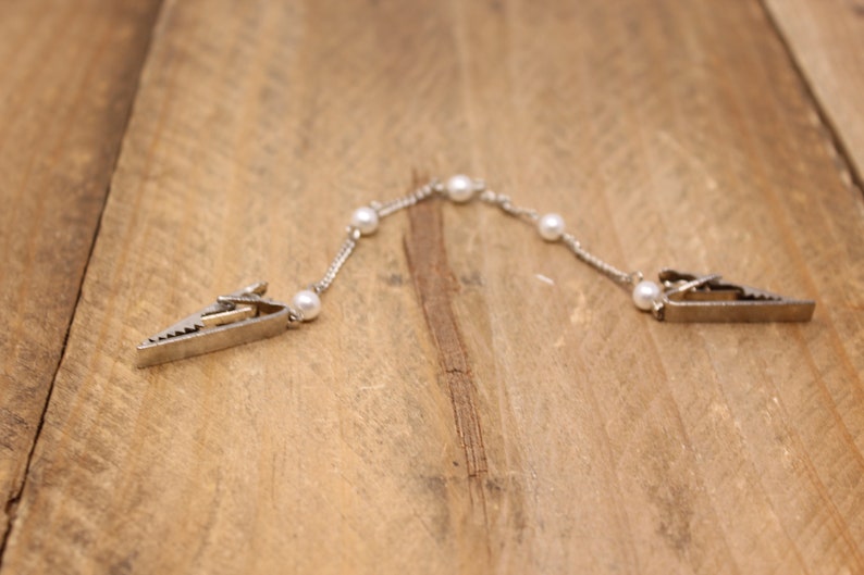 May include: A silver chain with white pearl accents and two silver clips on each end. The chain is lying on a wooden surface.