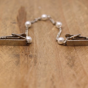 May include: A silver chain with white pearl accents connects two silver clips. The clips have a sawtooth edge and are designed to hold fabric together.