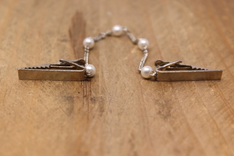 May include: A silver chain with white pearl accents connects two silver clips. The clips have a sawtooth edge and are designed to hold fabric together.