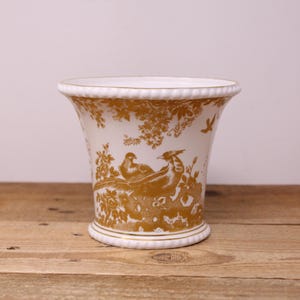 May include: A white ceramic vase featuring a gold-coloured transfer print of pheasants and floral foliage. The vessel has a flared rim and a base, both detailed with a raised beaded texture. The gold pattern wraps around the exterior surface.