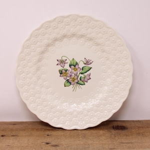 May include: A white ceramic plate with a scalloped edge and a floral design. The plate features a raised, textured pattern of small flowers around the rim. The center of the plate displays a bouquet of purple and yellow violets with green leaves.