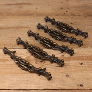 May include: Five antique bronze cabinet pulls with ornate, scrolling designs. Each pull features a central oval shape flanked by symmetrical, curved embellishments. The pulls are arranged diagonally on a wooden surface, showcasing their intricate details and aged finish.