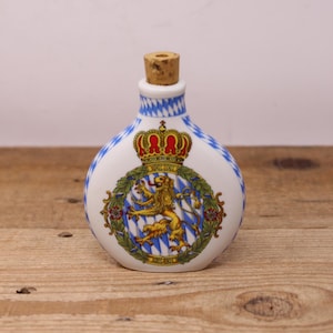 May include: A white ceramic bottle with a cork stopper. The bottle features a blue and white diamond pattern and a crest with a crown and a lion. The bottle is on a wooden surface.