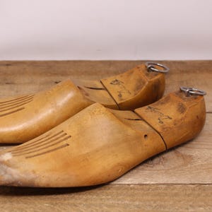 May include: A pair of light brown wooden shoe trees with a smooth finish, featuring a split-toe design with four parallel grooves on the forefoot, a hinged heel section, and a small metal pull ring attached to the back of each heel piece.