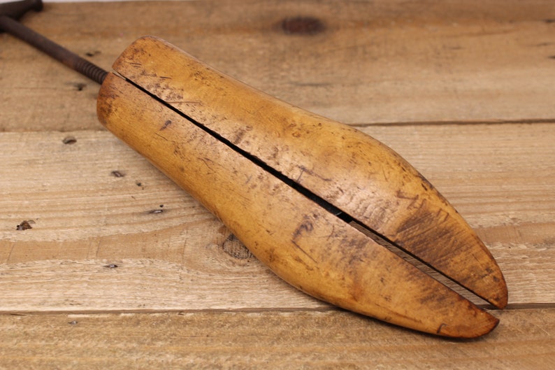 May include: A wooden shoe stretcher with a metal screw mechanism. The stretcher is made of light brown wood and has a split design to fit inside a shoe.