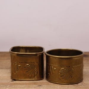 May include: Two antique brass napkin rings. Each ring has a cylindrical shape with a decorative embossed leaf design. The rings are made of a shiny, aged brass and sit on a wooden surface.