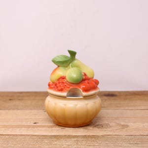 May include: A ceramic jar with a lid, featuring a fruit design. The lid is adorned with a cluster of red, yellow, and green fruits, including pears and berries. The jar has a light orange color and sits on a wooden surface.