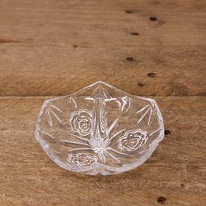 May include: A clear glass ring holder with a raised center post and a floral design. The holder is shaped like a flower with a scalloped edge.