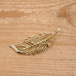May include: A gold-coloured, leaf-shaped brooch. The brooch features a detailed, openwork design with a smooth, curved edge and a textured, veined pattern. The brooch is on a light brown wooden surface.