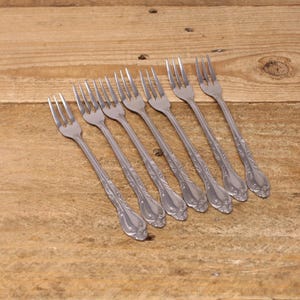 May include: Eight silver-coloured forks with decorative handles are arranged diagonally on a light brown wooden surface. The forks' tines and ornate patterns are visible. The forks are made of metal.