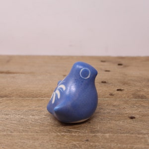 May include: A small, blue ceramic bird figurine with white decorative accents. The bird has a round body and a small beak. The figurine is sitting on a wooden surface. The bird's design is simple and elegant.
