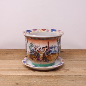 May include: A vintage ceramic planter with a matching saucer. The planter features a colourful, hand-painted scene of figures in a garden setting, with a blue, orange, and white floral border. The saucer has a matching design.