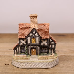 May include: A detailed miniature Tudor-style house with a red tiled roof, black and white timber framing, and a brick chimney. The house features a small arched doorway and a stone base, creating a charming, detailed miniature.