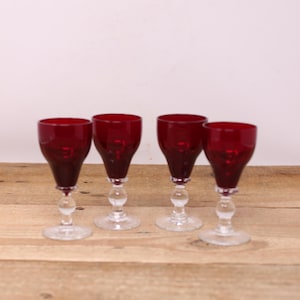 May include: Four antique ruby red wine glasses with clear, decorative stems and bases. The glasses are displayed on a wooden surface, highlighting their rich colour and elegant design. The stemware is suitable for serving wine.