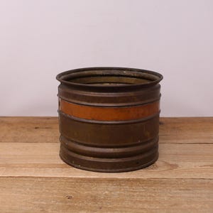 Peut inclure: Un récipient cylindrique en métal de couleur marron foncé, orné d'une bande horizontale orange-brun. Il repose sur une surface en bois, évoquant une esthétique vintage ou rustique. L'objet présente un aspect vieilli et patiné.