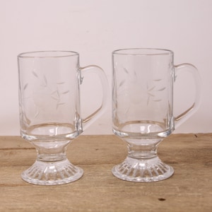 May include: Two clear glass mugs with etched designs and a footed base. The mugs have handles and are ideal for serving coffee or tea.