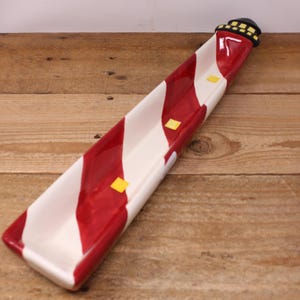 May include: A long, narrow ceramic tray shaped like a lighthouse, featuring diagonal red and white stripes. The top end includes a black lighthouse lantern room with yellow windows, and small yellow square accents are placed along the interior base.