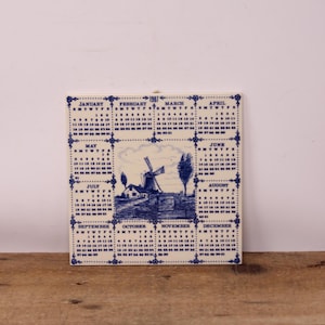 Peut inclure: Un calendrier carré en céramique pour l'année 1987, représentant un moulin à vent bleu et blanc. Le calendrier affiche les mois autour de l'image, avec la date de chaque jour clairement indiquée. La tuile a une bordure décorative.