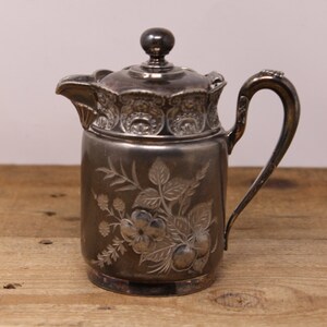 May include: A silver creamer with an ornate floral design. The creamer has a lid and a handle.
