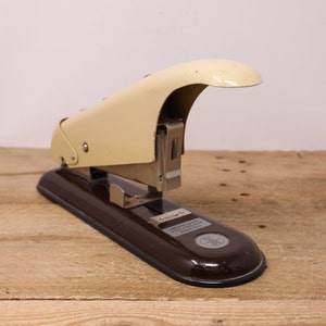 May include: A vintage brown and cream stapler with a metal base and a lever on the top. The stapler has a label that reads "Universal Stapler 2000 Series All Metal Parts Made in Germany".