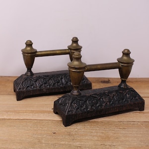 May include: A pair of antique fire dogs. The fire dogs have a dark brown, cast iron base with a floral design. The horizontal bar and finials are brass-coloured. They are displayed on a wooden surface.