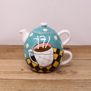 May include: A teal blue teapot with white polka dots and a white lid. The teapot has a white handle and spout. The teapot is decorated with a painted image of a coffee cup with steam rising from it. The coffee cup is on a black and yellow checkered background.