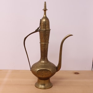 May include: A tall, antique-style brass ewer featuring an elongated neck, a curved handle, and a long, slender spout. The entire surface of the metal vessel is covered in intricate, etched floral patterns and decorative engravings throughout.