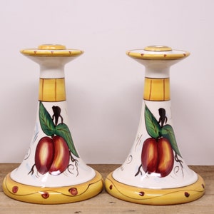May include: A pair of white ceramic candle holders with a yellow, red and green fruit design. The holders are tall and slender with a wide base.
