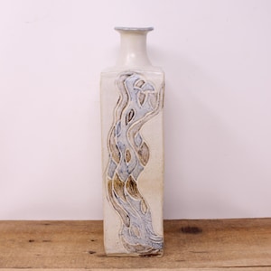 May include: A tall, rectangular ceramic vase with a cream-colored base and a light blue and brown abstract design. The vase has a narrow neck and a slightly flared rim. The design resembles a flowing river or stream.