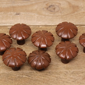 May include: Eight brown, flower-shaped cabinet knobs. Each knob has a detailed, carved design on the top and a short, cylindrical stem. The knobs are arranged on a light brown wooden surface.