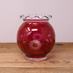 May include: A round, deep red glass vase featuring a clear, ruffled top rim and a clear base. The body of the vase is decorated with an etched floral pattern consisting of two prominent flowers and delicate leaves encircling the centre.