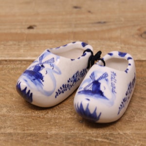 May include: Two blue and white ceramic Dutch clogs with a windmill design. The clogs are tied together with a blue string. The clog on the right has the word "Holland" written on it.