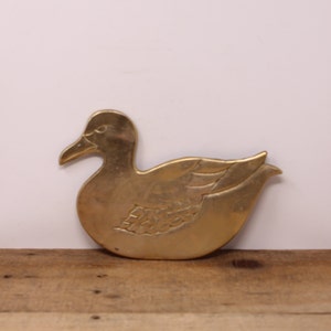 May include: A gold-coloured metal duck-shaped trivet. The duck is sitting on a wooden surface.