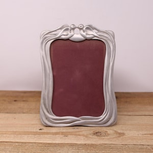May include: A silver-coloured picture frame with a decorative, flowing design. The top of the frame features two stylised birds. The frame holds a burgundy-coloured insert and is displayed on a wooden surface.