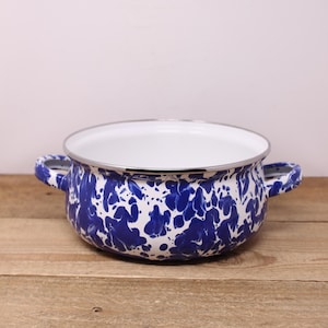May include: A white enamel Dutch oven with blue marbling and silver trim. The oven has two handles and is sitting on a wooden surface.