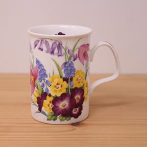 May include: A white ceramic mug featuring a colorful botanical illustration of purple bluebells, blue grape hyacinths, yellow primroses, and deep purple flowers with green leaves. The mug has a smooth cylindrical body and a curved handle on the side.
