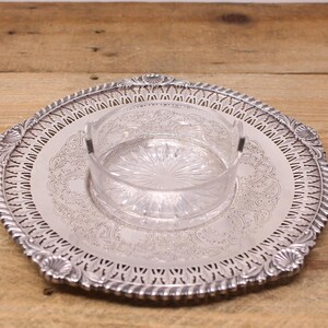 May include: A silver serving tray with an engraved pattern and a clear glass bowl. The tray has a decorative edge with cut-out patterns. The bowl sits in the centre of the tray and has a starburst pattern on the base.