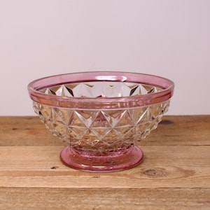 May include: A clear glass bowl featuring a diamond-cut geometric pattern across its exterior. The bowl has a circular base and a wide rim, both finished with a vibrant pink tint that contrasts with the transparent body of the glass vessel.