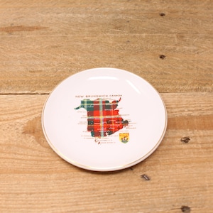 May include: A white ceramic plate with a gold rim. The plate features a map of New Brunswick, Canada, in a plaid pattern with the text "NEW BRUNSWICK CANADA". A decorative item for the home.