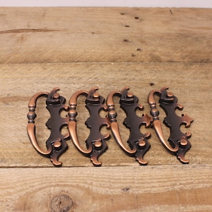 May include: Four antique bronze cabinet pulls with a decorative design. Each pull features a curved handle and ornate backplate. The pulls are arranged on a wooden surface, showcasing their vintage style and potential for furniture restoration.