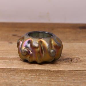 May include: An iridescent glass bowl with a ruffled, organic shape. The bowl has a dark interior and a shimmering exterior with hues of gold, purple, and green. The bowl sits on a wooden surface.