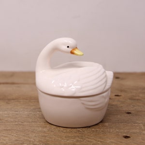 May include: White ceramic swan-shaped trinket box with a yellow beak and black eyes. The box is closed and the swan's head is turned to the side.