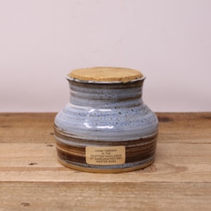 May include: A hand-thrown ceramic jar with a light blue and brown glaze, topped with a tan cork lid. The jar has a rounded shape and a label that reads "Hand Thrown in the Scottish Highlands at Gairloch Pottery Wester Ross."