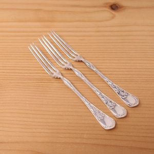 May include: Three silver-coloured metal forks arranged diagonally. Each fork features a four-tine head and a slender handle decorated with an embossed floral pattern that extends from the neck down to the rounded, smooth end of the handle.