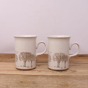 May include: Two beige ceramic mugs with a brown tree and bird design. The mugs have a curved handle and a slightly flared rim. The mugs are on a wooden surface.