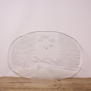 May include: A clear glass serving platter with a raised design of a countryside scene with birds flying over a small town and clouds. The platter has a textured surface.