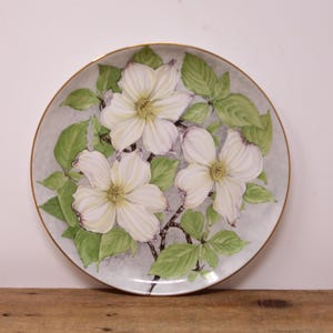 May include: A round decorative plate with a floral design. The plate has a gold rim and a light grey background. Three white dogwood flowers with yellow centres are painted on the plate, surrounded by green leaves and brown stems.