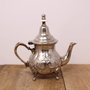 May include: A silver teapot with ornate floral designs and a curved handle. The teapot has a small spout and sits on three small feet. The lid is topped with a white finial. The teapot is made of metal.